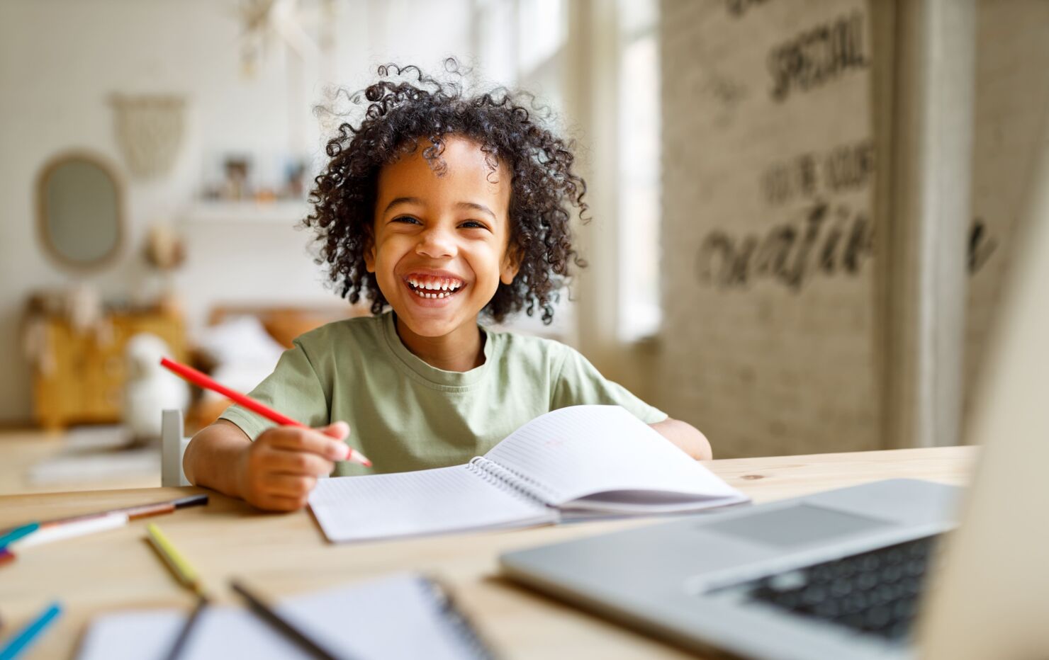 Ein lachendes Kind, welches an einem Tisch sitzt und auf einem Notizheft mit einem roten Stift vor einem Laptop abmalt.