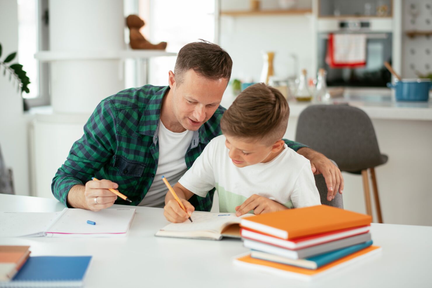 Ein Vater sitzt mit seinem Sohn an einem Tisch und hilft ihm die Hausaufgaben zu erledigen. Er hat mehrere Bücher vor sich.