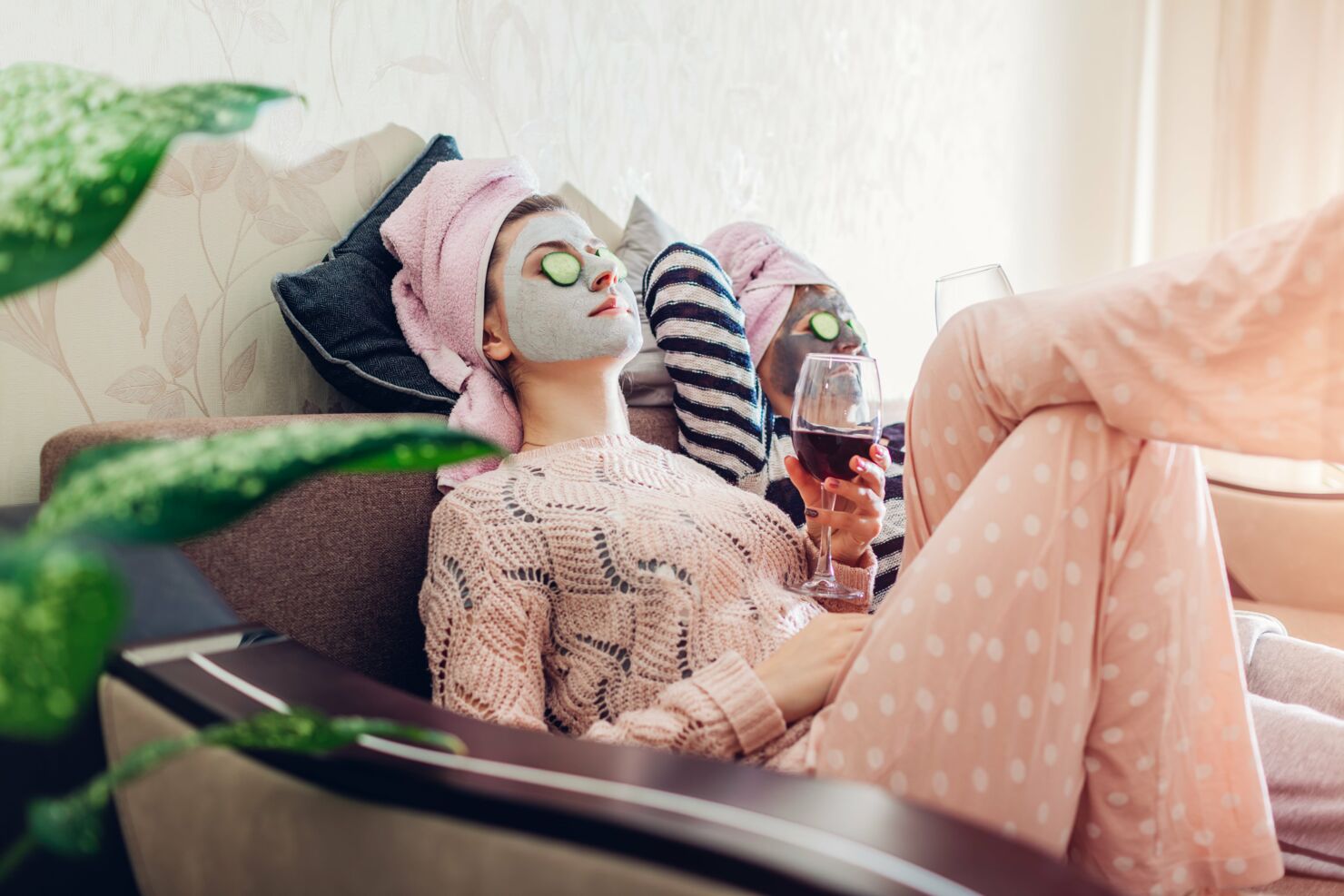 Eine erwachsene junge Frau trägt eine Maske und hat Gurken auf den Augen, trinkt nebenbei Wein mit der Kollegin