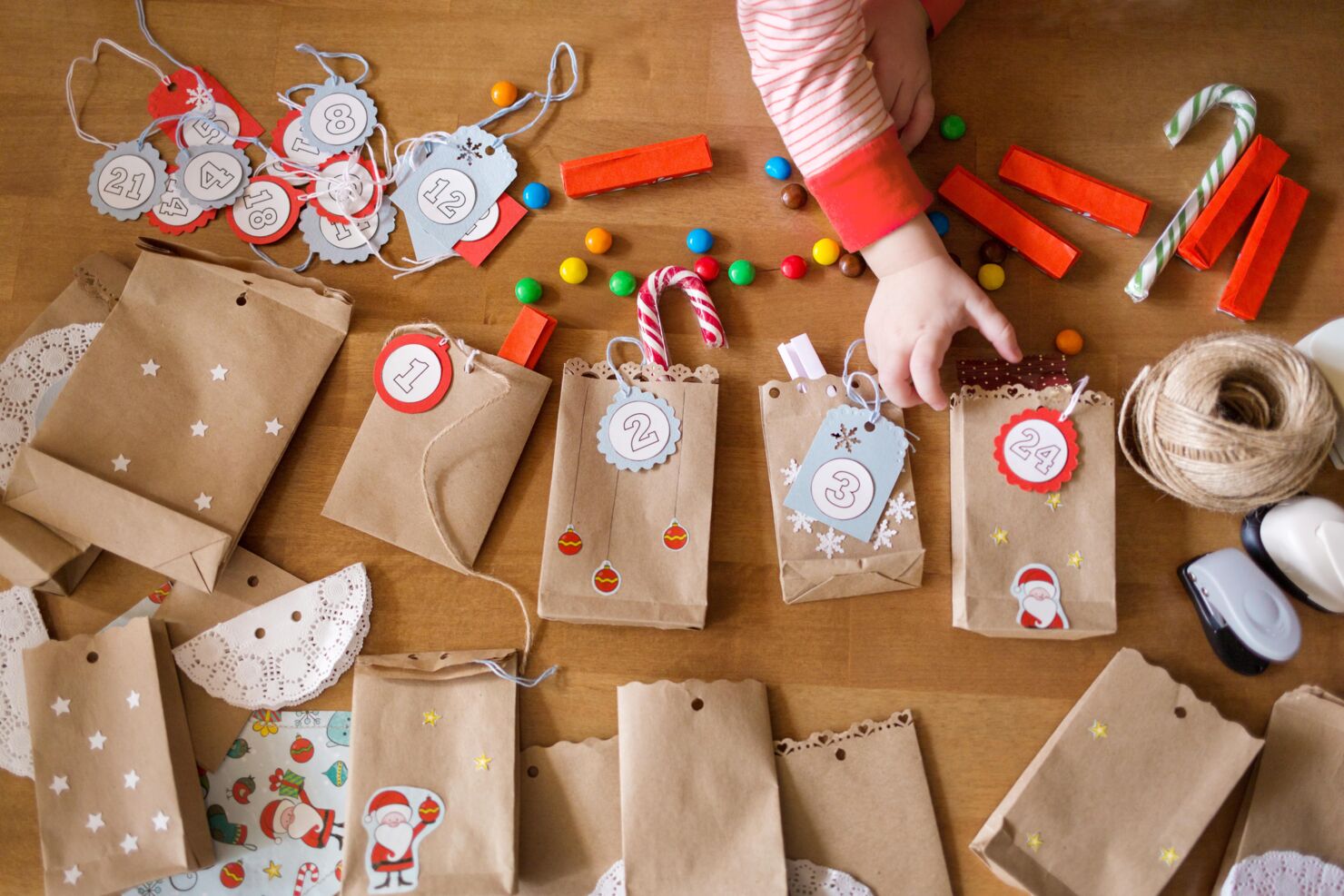 Eine Kind macht ein DiY Adventskalender, die Sterne und einen Weihnachtsmann-Sticker drauf hat. Es hat M&M's und mehrere Papiertaschen auf dem Boden